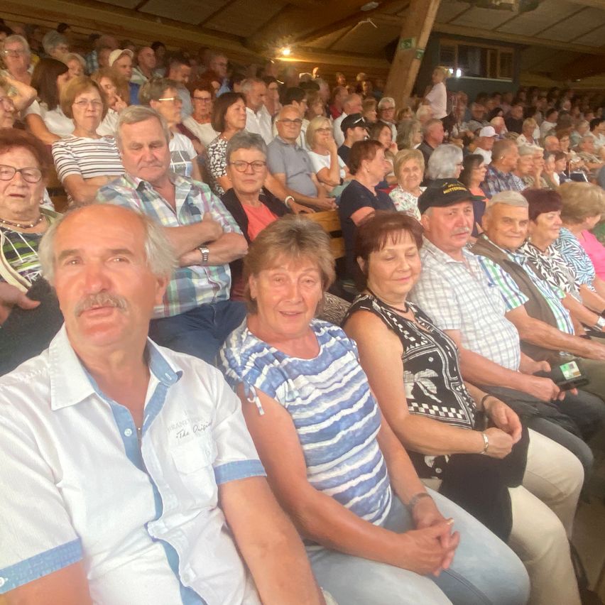 Eine Gruppe älterer Erwachsener sitzt in einem Stadion, einige tragen Brillen und Hüte. Sie scheinen eine Veranstaltung zu verfolgen.