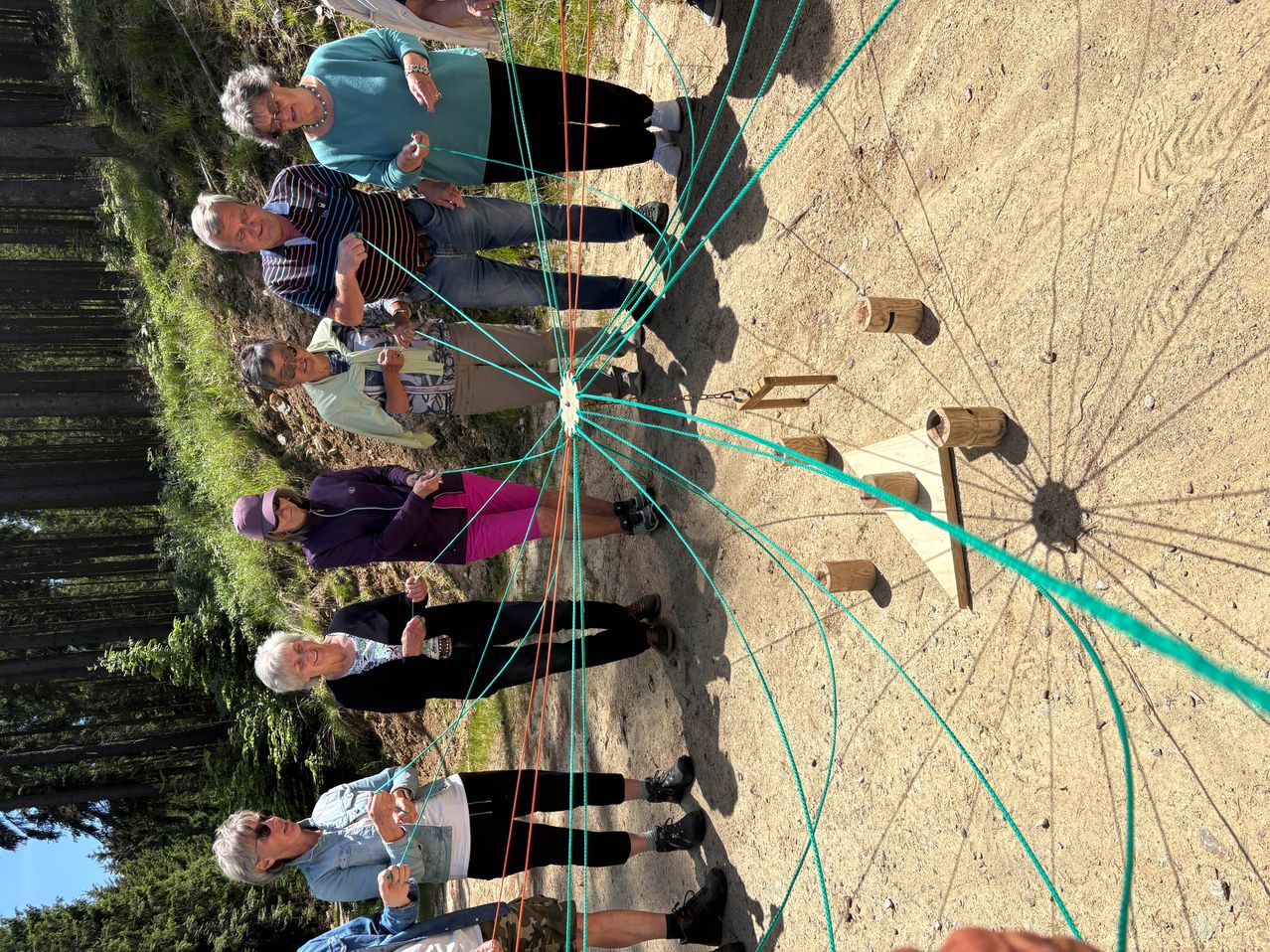 A group of people are holding colored ropes and standing in a circle, forming a wheel. The setting is outdoors with dirt ground and some trees.