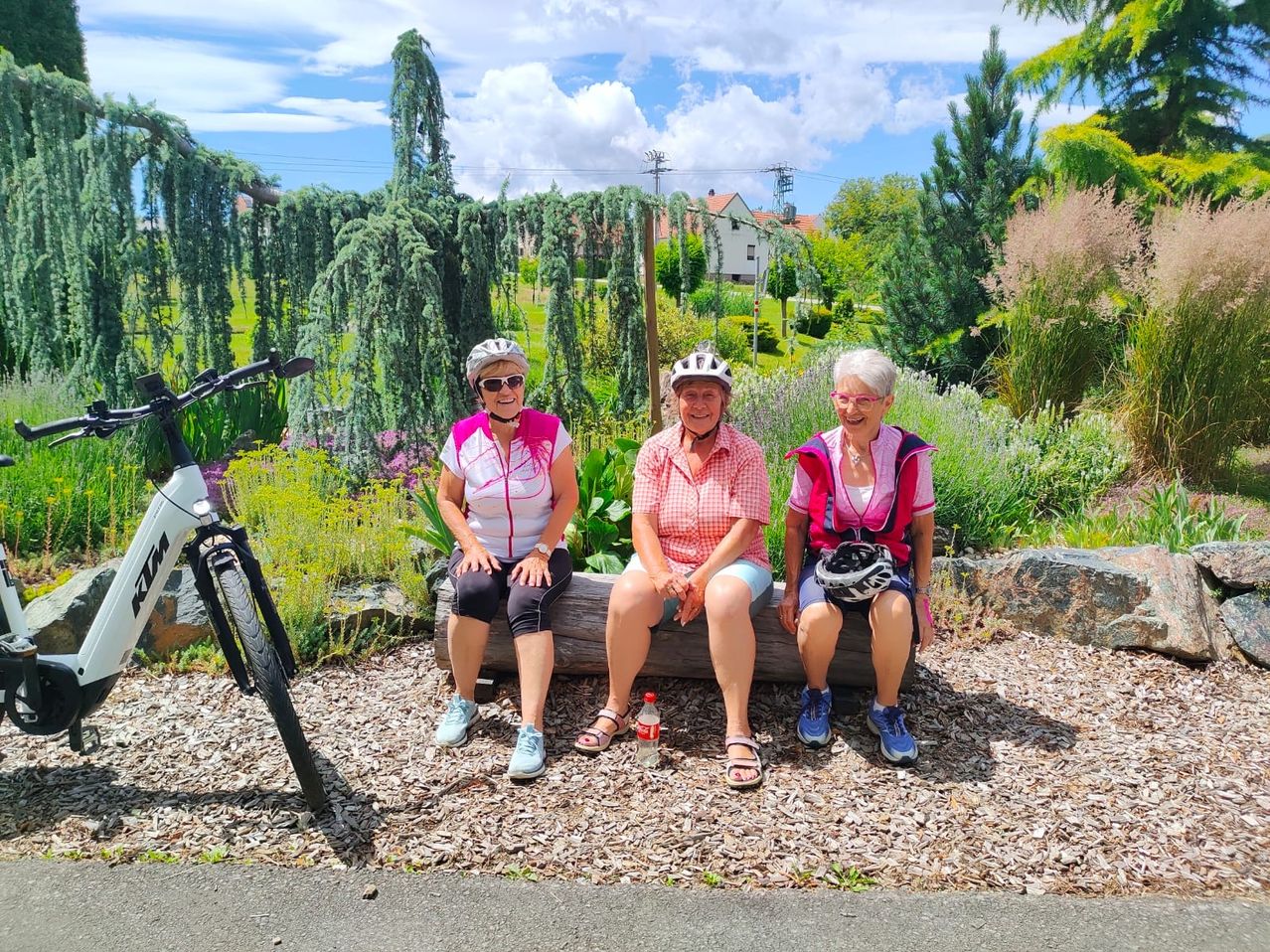 Drei Frauen in Fahrradkleidung sitzen auf einem Holzstamm, lächeln und posieren für ein Foto. Ein Fahrrad ist in der Nähe geparkt. Sie sind von einem Garten mit Pflanzen und Bäumen umgeben.