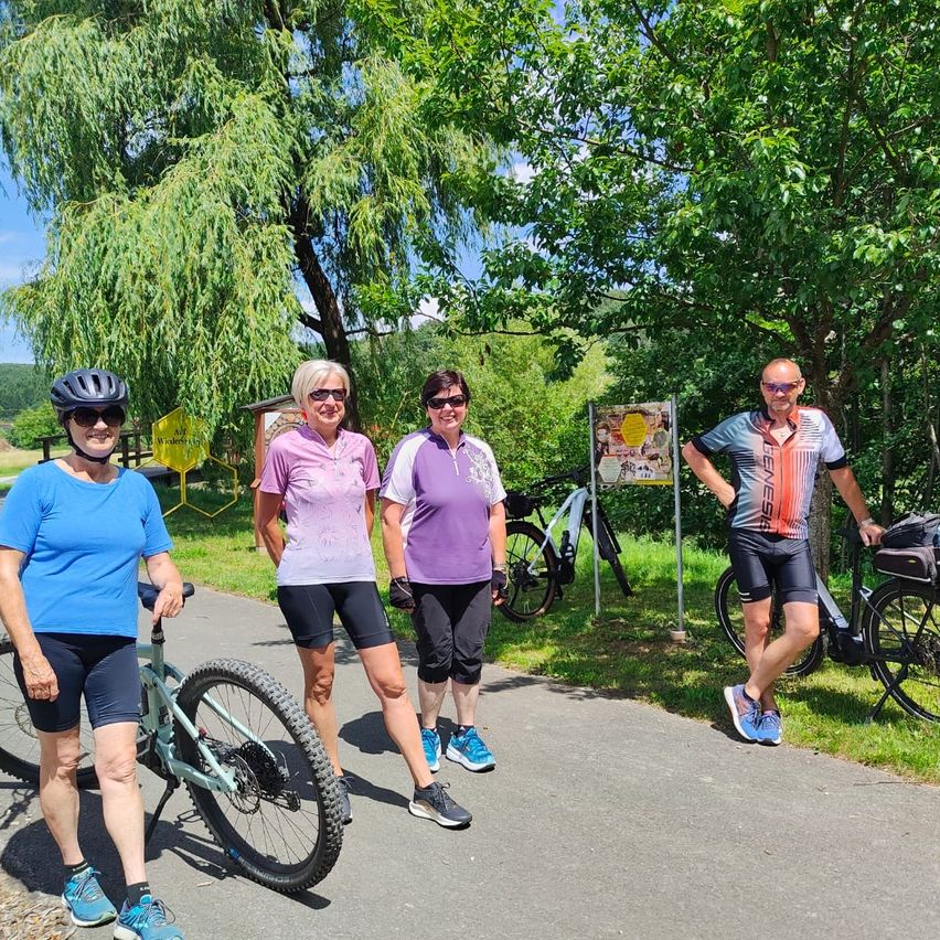 Vier Radfahrer posieren auf einem Pfad, umgeben von üppiger Grünfläche und Bäumen, für ein Foto. Jeder trägt einen Helm und Radfahrerkleidung. Ein Schild und ein Fahrrad sind im Hintergrund.