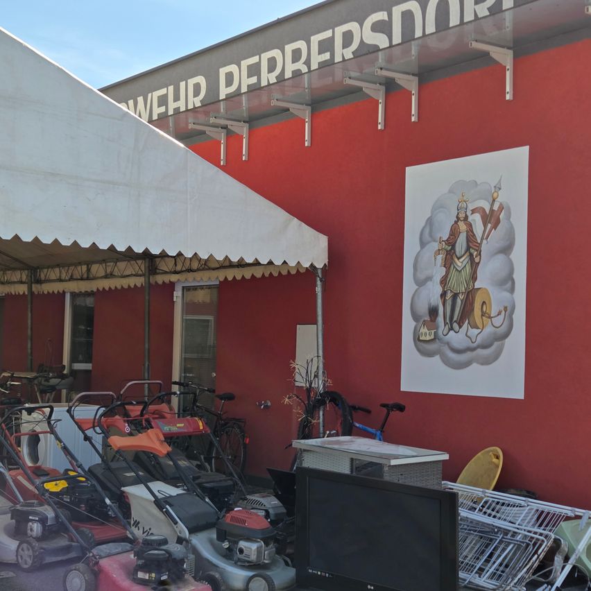 A red building with 'WEHR PERFERSMORT' written above. A large poster is attached to the wall. Many items, including lawn mowers and bicycles, are kept in front.