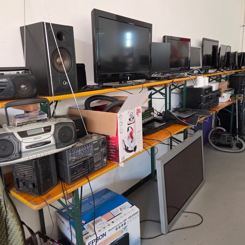 A cluttered room filled with electronic devices, including monitors, speakers, a vacuum cleaner, and a boombox, placed on shelves.