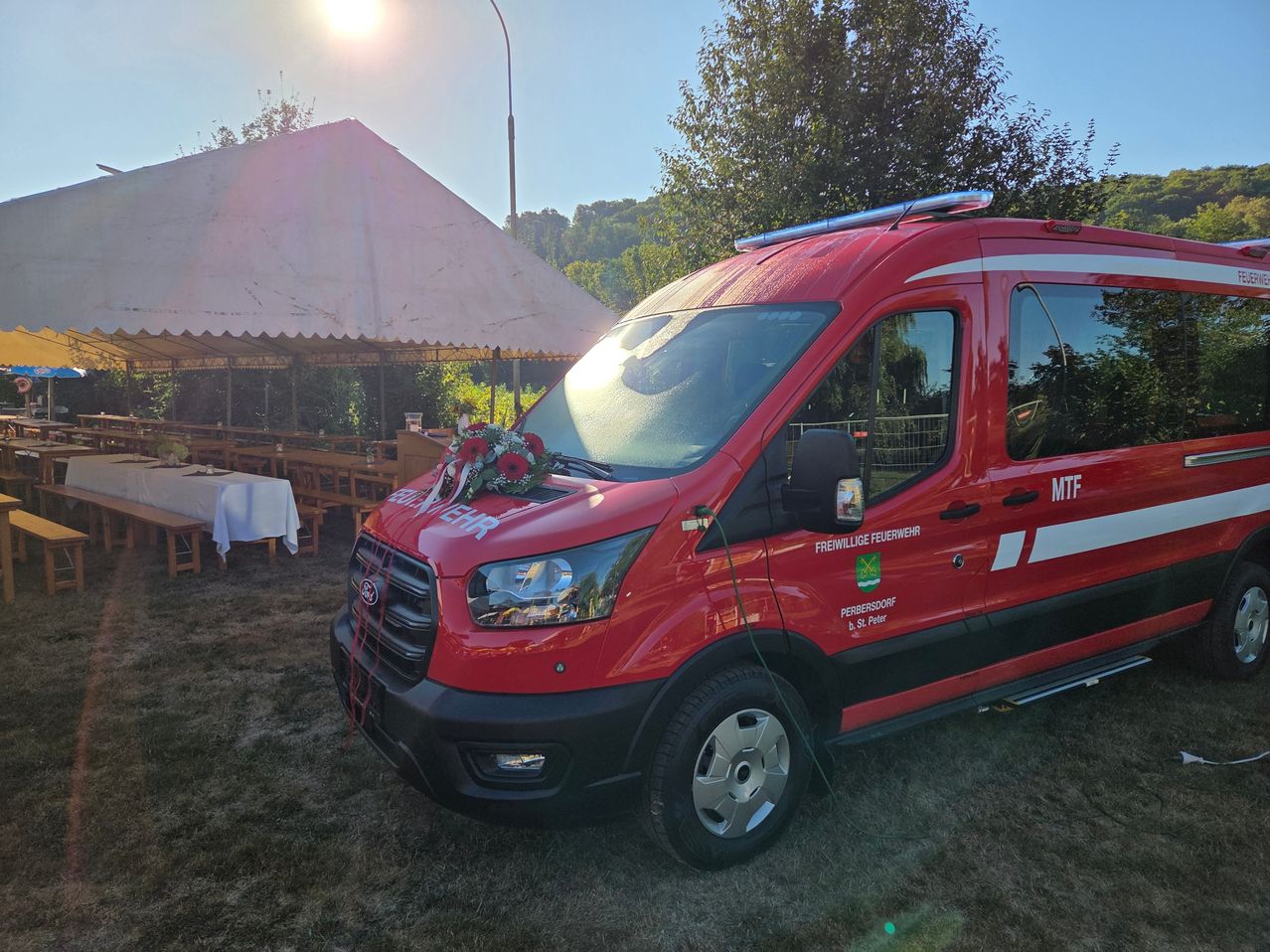 A red fire truck with a bouquet of flowers on the front hood is parked in a grassy area with a tent and tables in the background.