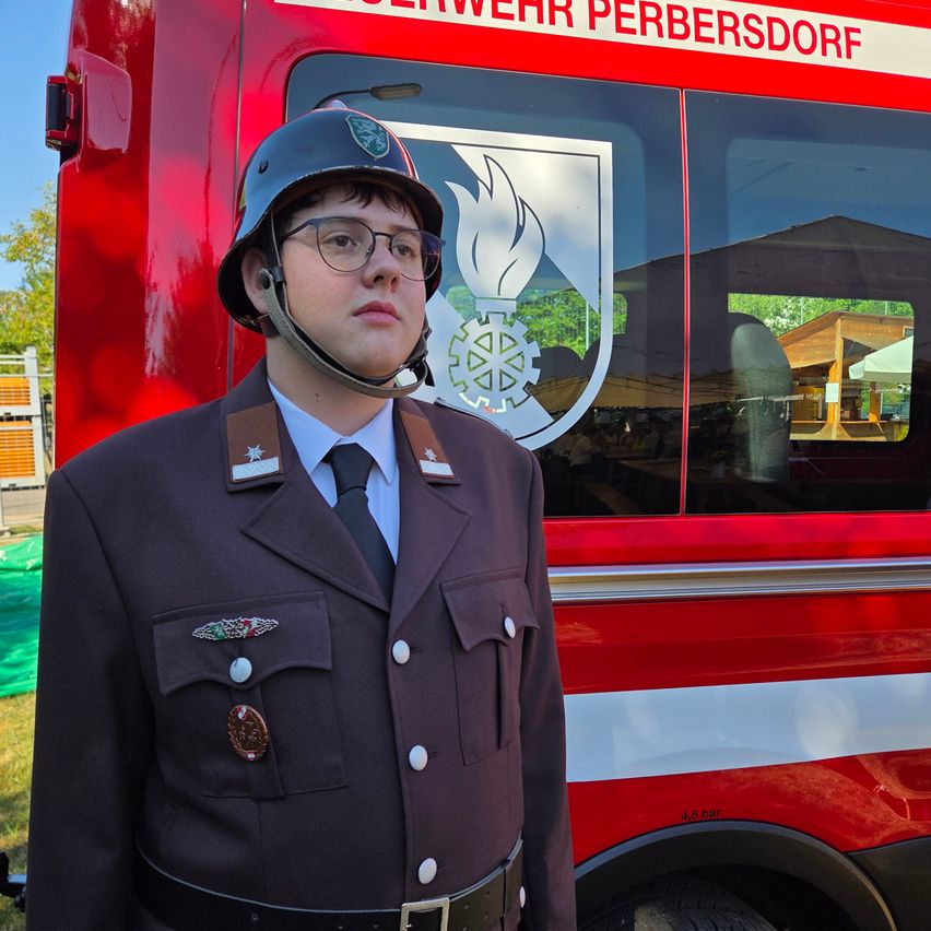 A firefighter in uniform stands in front of a red fire truck with a logo on the side.