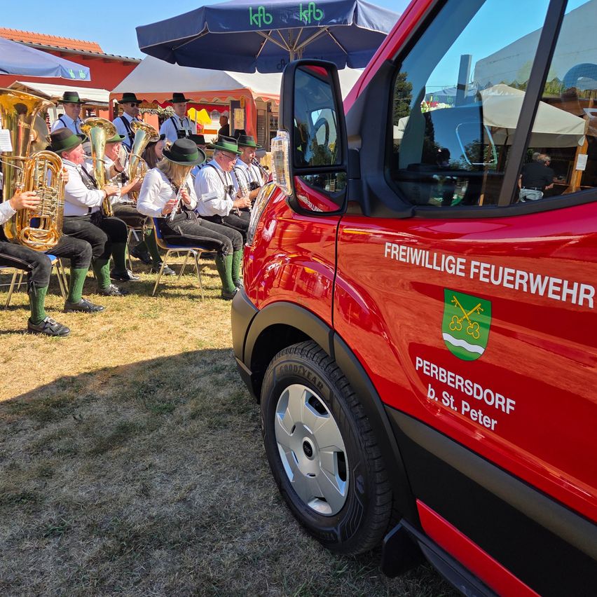 A group of musicians in traditional attire playing brass instruments under an umbrella at an outdoor event. A red fire truck is parked nearby.