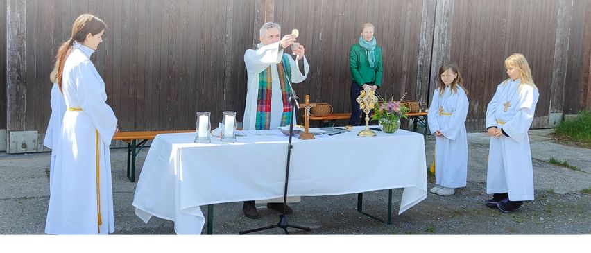 Ein Priester steht an einem Altar und hält Kelch und Patene. Zwei Kinder und eine Frau in Grün stehen in der Nähe. Der Altar ist mit Kerzen, Blumen und einem Kreuz geschmückt. Der Textüberlagerung lautet 'Christi Himmelfahrt gemeinsame Feldmesse im Pfarrverband bei der Flughalle - Eucharistiefeier'.