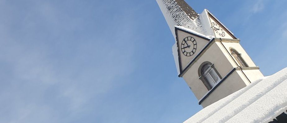 Ein Gebäude mit einem Schnee bedeckten Glockenturm, einem Schornstein und mehreren Glasfenstern, mit einem verschneiten Baum im Hintergrund und einem blauen Himmel.