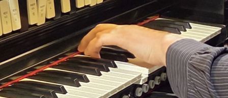 A person's hand is pressing keys on a large black and white organ. The organ has a row of white switches above the keys.