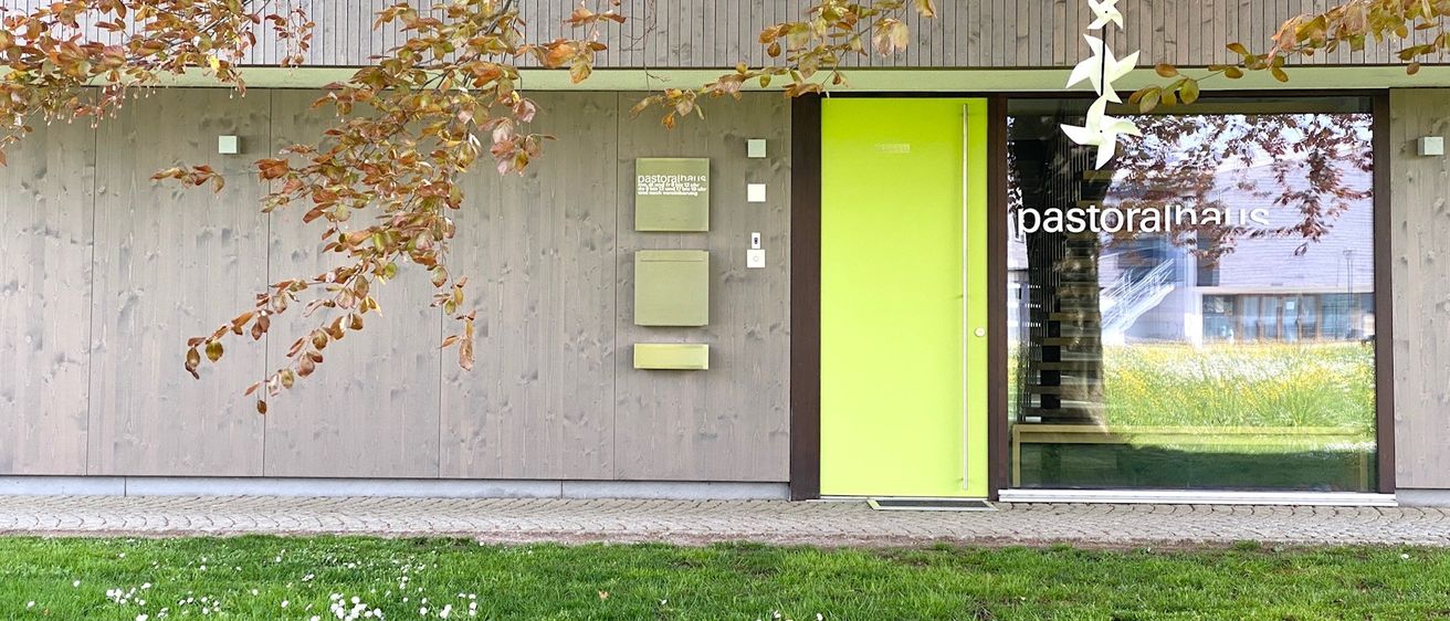 A building exterior with a bright yellow door and three green mailboxes with the text 'pastoralhaus' on the wall.