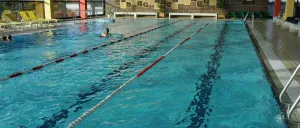 Ein Hallenschwimmbad mit durch rote und weisse Seile markierten Bahnen. Ein Schwimmer schwimmt in einer der Bahnen. Das Becken ist von einer dekorativen Wand und Bänken umgeben.