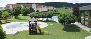 Ein Luftbild eines Resorts mit einem Swimmingpool, einem Spielplatz und Gebäuden, umgeben von üppiger Grünfläche und Bergen in der Ferne.