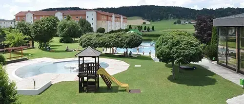 Ein Luftbild eines Resorts mit einem Swimmingpool, einem Spielplatz und Gebäuden, umgeben von üppiger Grünfläche und Bergen in der Ferne.