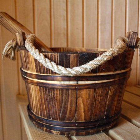 Ein Holzeimer mit einem Seilgriff steht auf einer Holzbank. Der Eimer ist leer und hat einen kleinen Holzstab darin.