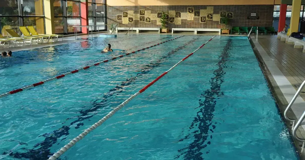 Ein Hallenschwimmbad mit einem Schwimmer, durch rote und weiße Bojen markierte Bahnen und dekorative Fliesen an den Wänden.