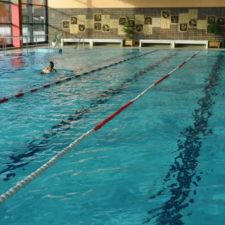 Ein Hallenschwimmbad mit einem Schwimmer, durch rote und weiße Bojen markierte Bahnen und dekorative Fliesen an den Wänden.