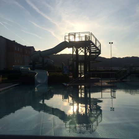 Ein Wasserrutschenpark in der Nähe eines Gebäudes mit Bergen im Hintergrund bei Sonnenuntergang. Die Sonne spiegelt sich im Wasser.