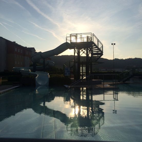 Ein Wasserrutschenpark in der Nähe eines Gebäudes mit Bergen im Hintergrund bei Sonnenuntergang. Die Sonne spiegelt sich im Wasser.