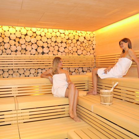 Zwei Frauen in einer Sauna, in Handtücher gewickelt, entspannen auf Holzbänken mit einem hölzernen Eimer in der Nähe. Der Raum ist warm, beleuchtet durch weiches gelbes Licht.