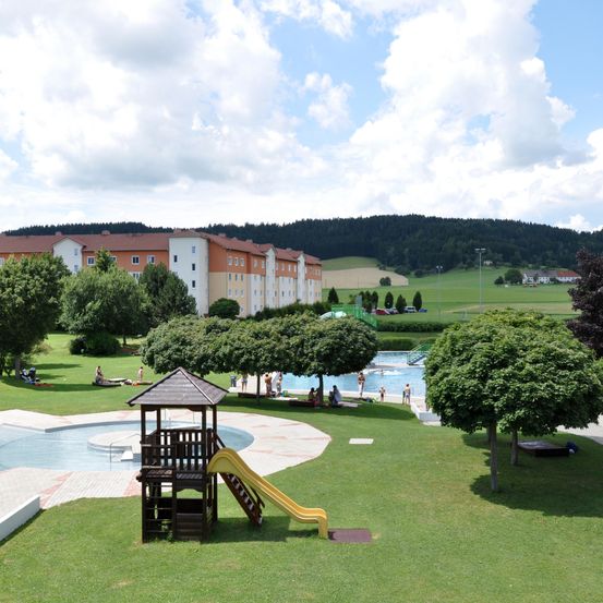 Luftaufnahme eines Erholungsgebiets mit zwei Pools, einem mit einer Rutsche, umgeben von Bäumen und Gras. Im Hintergrund befindet sich ein Gebäude mit mehreren Fenstern. Menschen schwimmen und entspannen sich auf der Wiese.