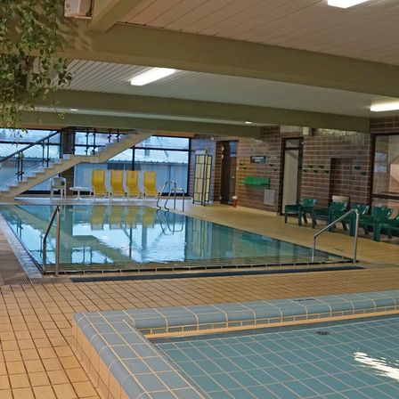 Ein Hallenbad mit gelben Stühlen und grünen Bänken, beleuchtet von Deckenleuchten, und einer Treppe, die zu einer Glastür führt.