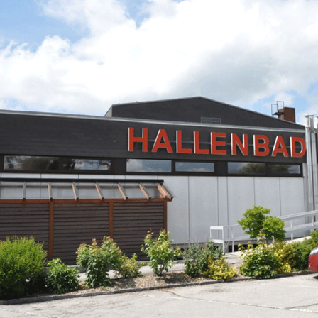 Ein rotes Auto ist vor einem Gebäude mit dem Wort Hallenbad geparkt. Das Gebäude hat eine Treppe und eine Glastür.