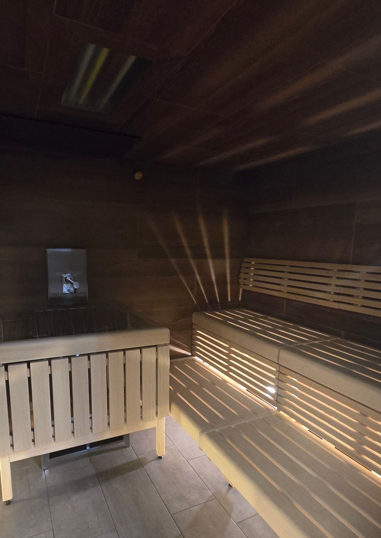 Ein leerer Holzsaunaraum mit Holzbänken und einer Holzwand. Der Raum wird von einem Licht an der Decke beleuchtet.