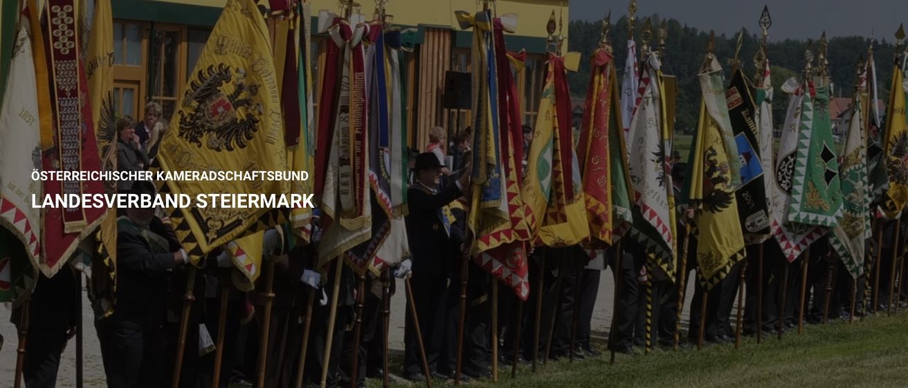 Eine Gruppe von Menschen steht vor mehreren Fahnen, die an Stangen befestigt sind, auf einer Rasenfläche.