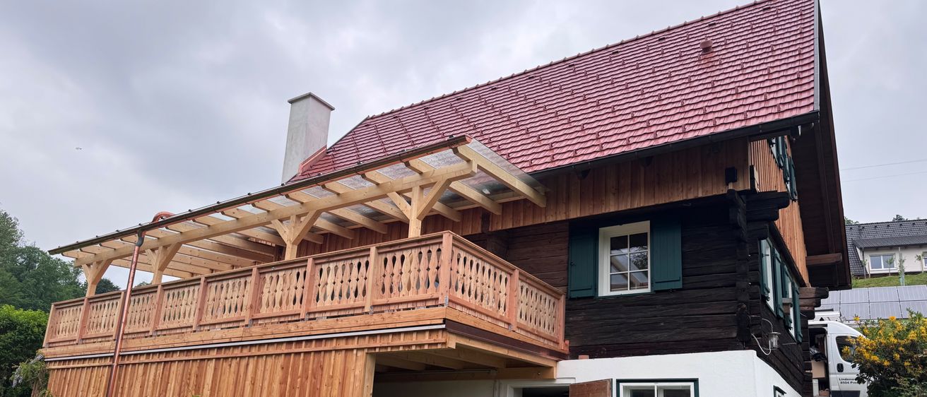 Ein traditionelles Holzhaus mit rotem Ziegeldach und Holzbalkon. Der Eingang ist offen mit einer Holztür und einem kleinen Baumstumpf in der Nähe.