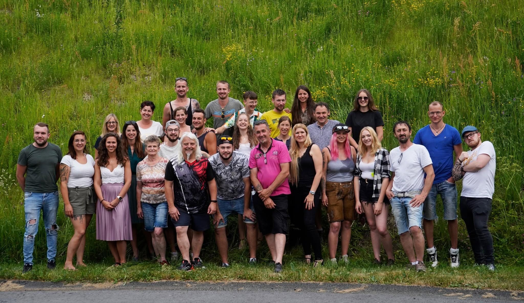 Eine Gruppe von Menschen, darunter Männer und Frauen, posiert für ein Foto in einem Grasbereich. Sie alle lächeln und scheinen sich zu amüsieren.