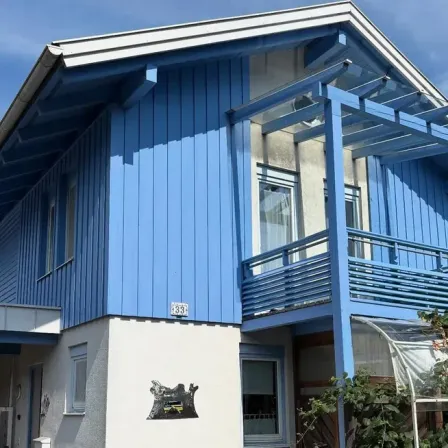 Ein modernes blaues Haus mit weißen Verkleidungen und einem Balkon, ausgestattet mit einem Glasgewächshaus und einem schwarzen Emblem an der Wand.