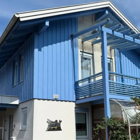 Ein modernes blaues Haus mit weißen Verkleidungen und einem Balkon, ausgestattet mit einem Glasgewächshaus und einem schwarzen Emblem an der Wand.