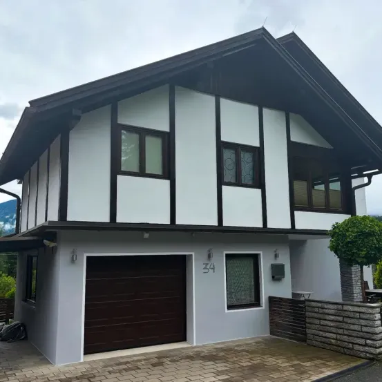 Ein zweistöckiges Haus mit einer braunen Garagentür und weißen Wänden, mit einem schwarzen Dach und einem kleinen Baum davor.