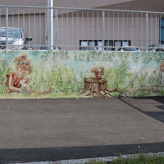 Ein großes Wandbild zeigt ein Mädchen, das in einem Feld mit einem Hasen und einem Eichhörnchen sitzt. Die Wand befindet sich neben einem Parkplatz mit geparkten Autos.