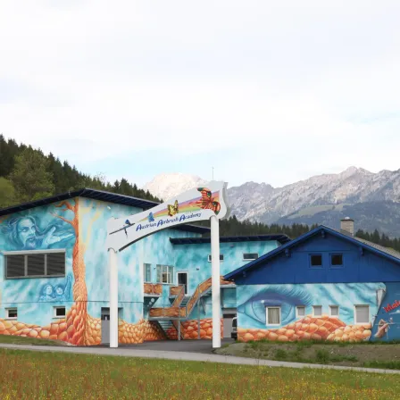 Ein blaues Gebäude mit farbenfrohen Wandgemälden und einem Schild, das Austrian Airbrush Academy vor einer Bergkette liest.
