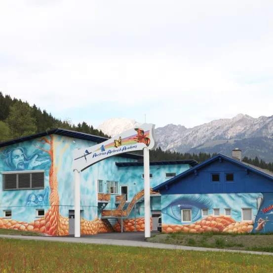 Ein blaues Gebäude mit farbenfrohen Wandgemälden und einem Schild, das Austrian Airbrush Academy vor einer Bergkette liest.