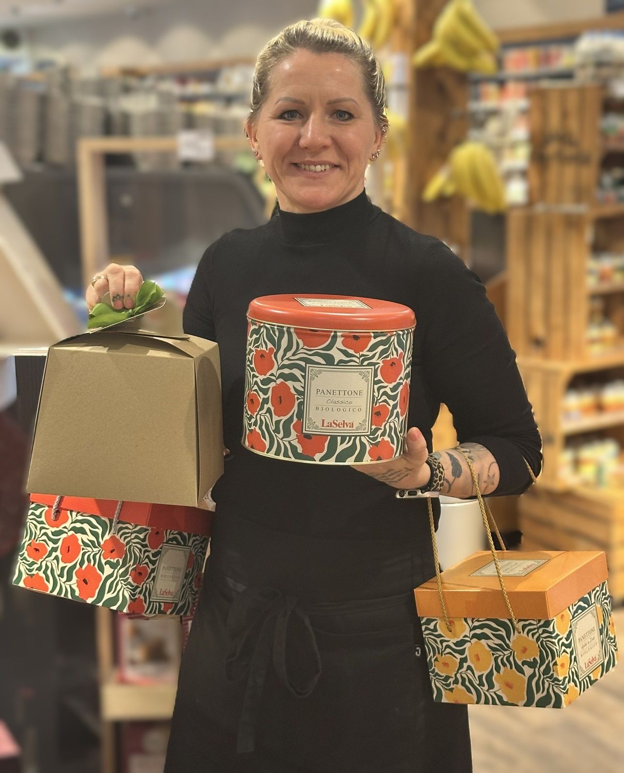 Eine Frau in einem schwarzen Rollkragenpullover steht in einem Geschäft und hält eine rote zylindrische Dose mit der Aufschrift 'Panettone Classico Biologico' und zwei Blumengeschenkboxen.