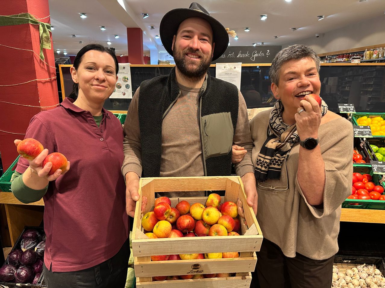Three people pose for a photo in a market. A man holds a crate of apples, a woman on his left holds an apple, and a woman on his right eats an apple. Behind them is a blackboard with text and a red pillar.