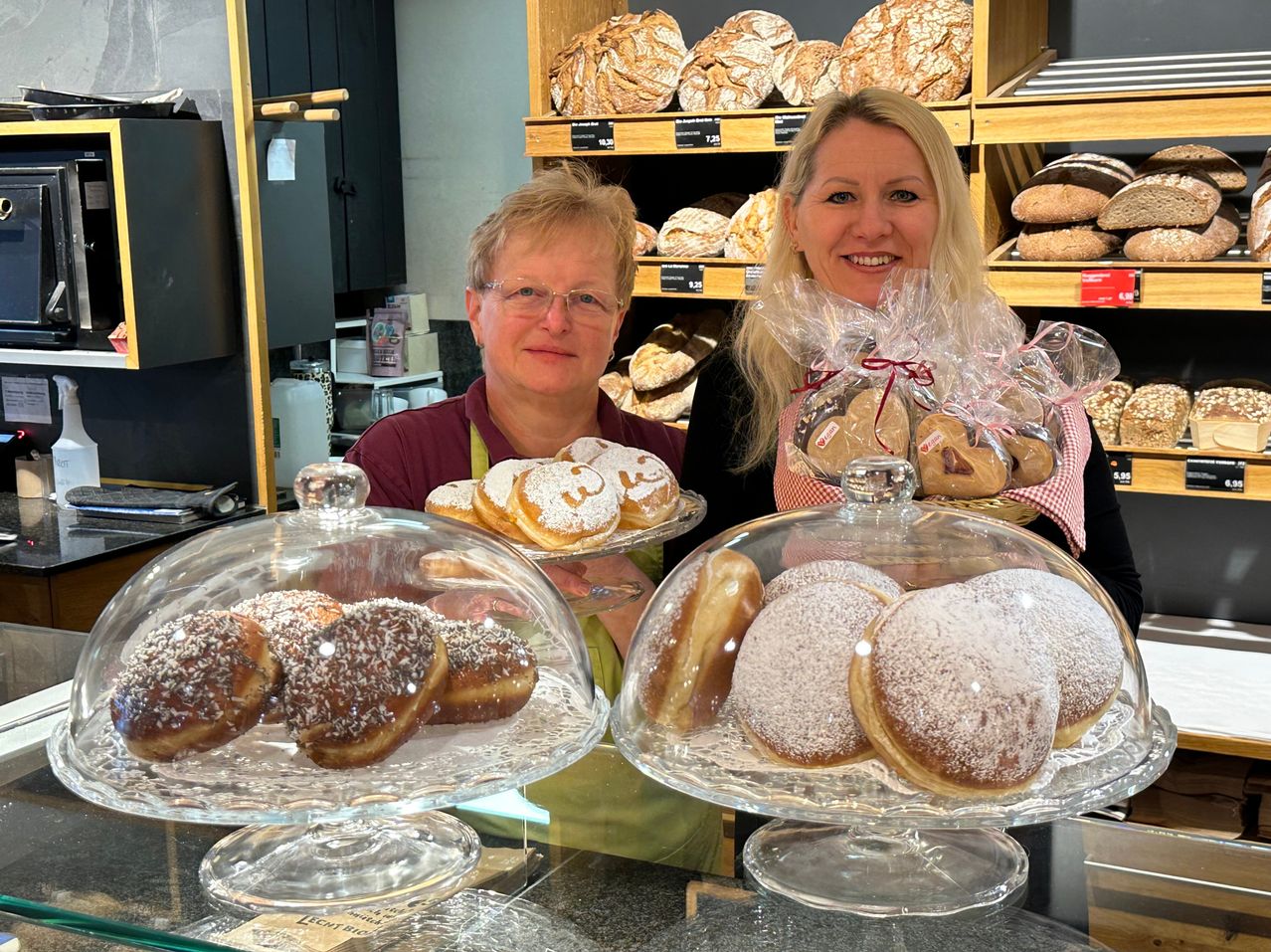 Two women stand behind a counter in a bakery, smiling. One holds a tray of powdered donuts, and the other has a basket of heart-shaped cookies. Behind them are shelves of bread.