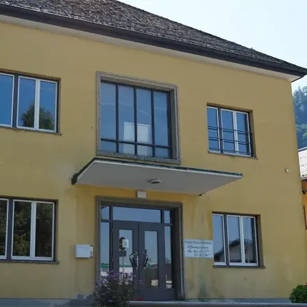 Ein gelbes Gebäude mit mehreren Fenstern und einer Glastür, mit einem kleinen Vordach. Über der Tür befindet sich ein Schild mit Text und Zahlen.