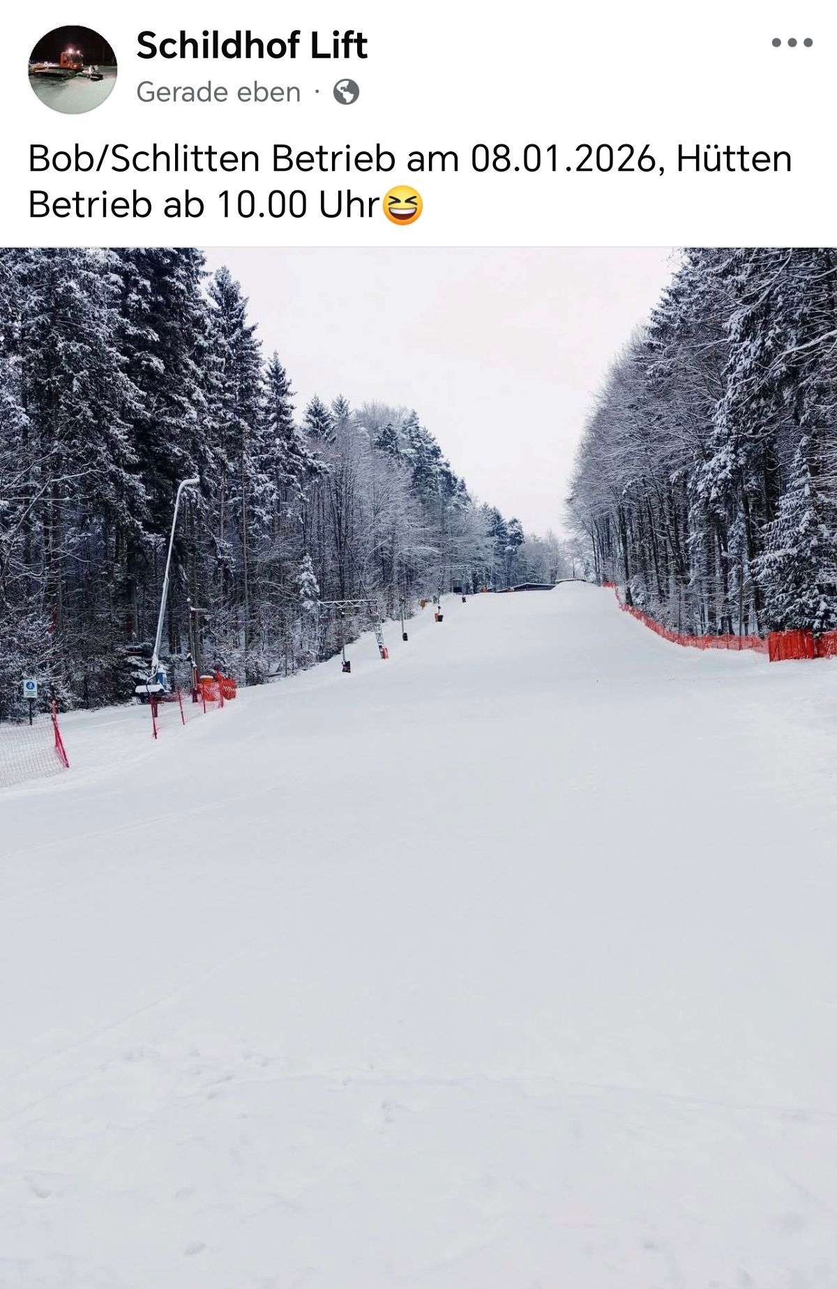 Eine verschneite Skipiste mit Tannenbäumen auf beiden Seiten. Die Piste hat eine glatte, kompakte Oberfläche, und orange Sicherheitsbarrieren markieren die Ränder. Ein Straßenlicht steht auf der linken Seite.
