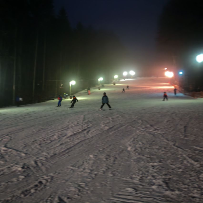 Several people are skiing down a snowy slope at night, illuminated by streetlights. The sky is dark, and there are trees around the slope.