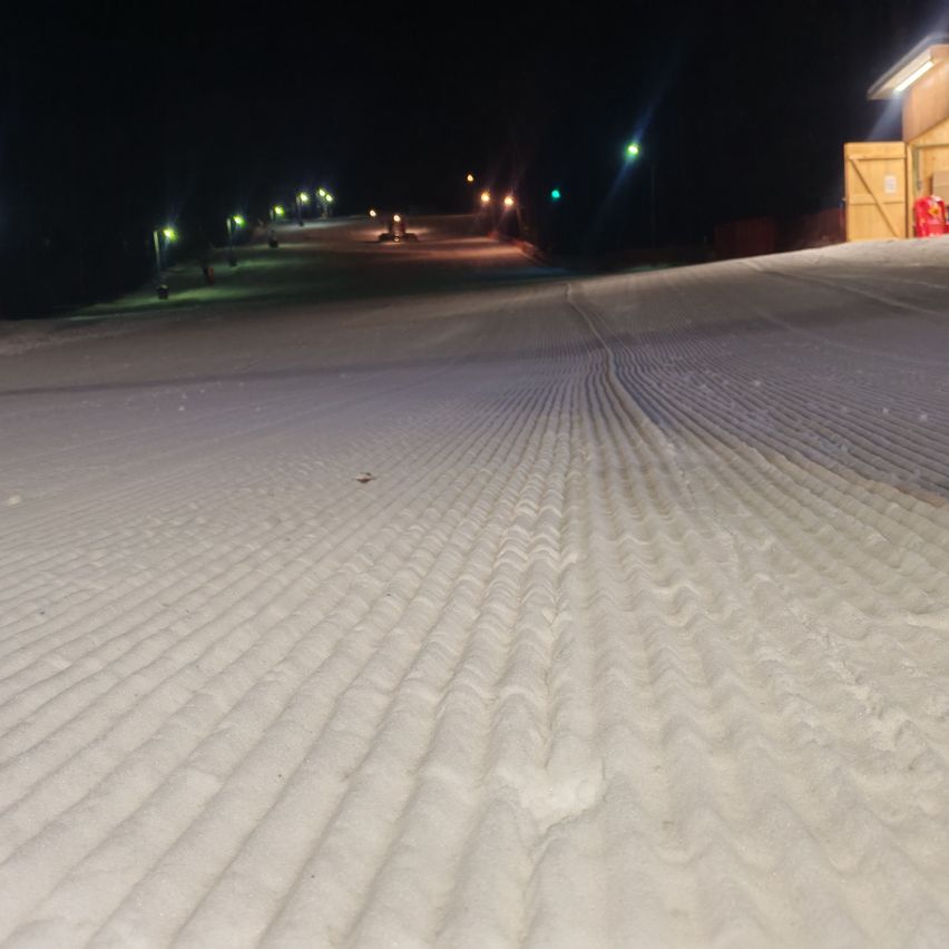 A night view of a snow-covered slope with lights illuminating the path. A building is visible in the background.