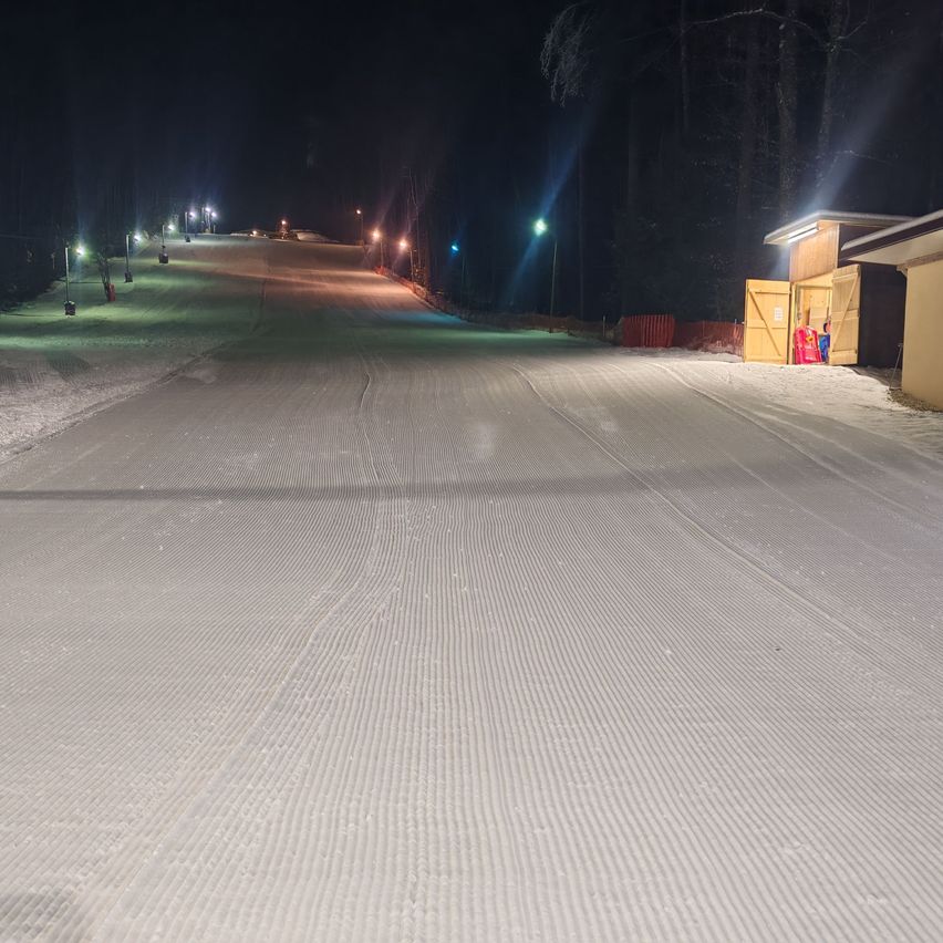 A snowy slope at night is illuminated by streetlights. The area is surrounded by trees, and there is a building with an open door.