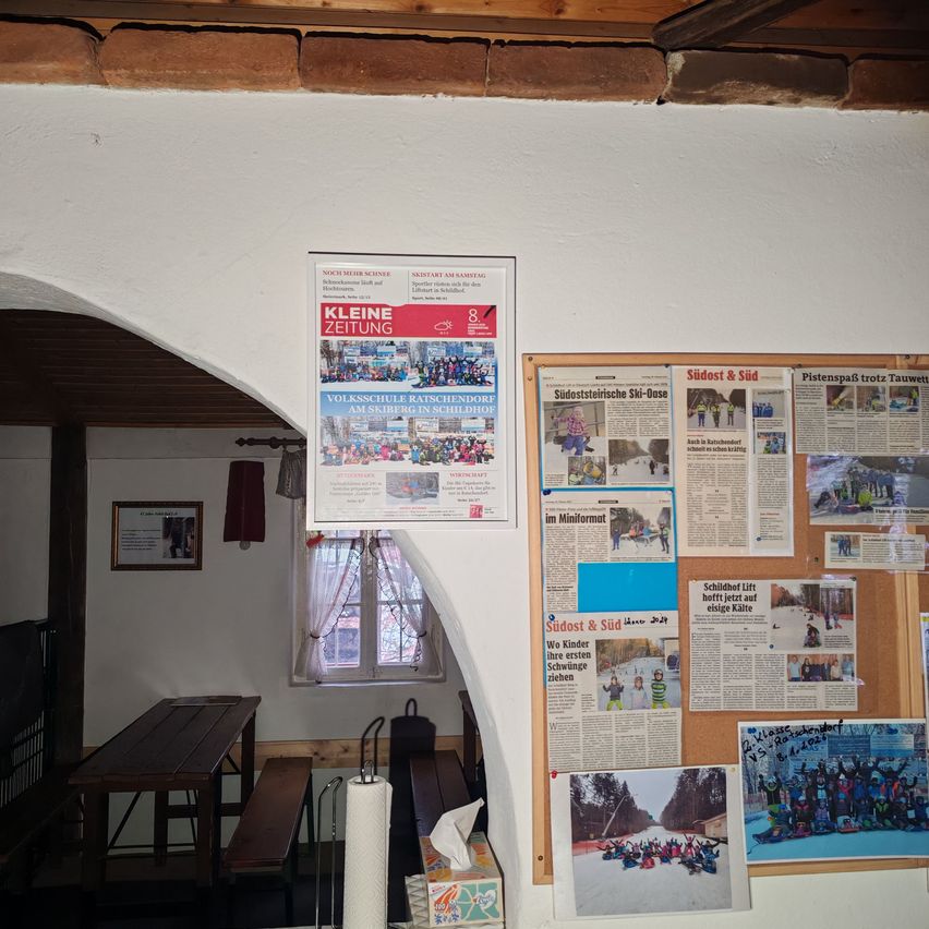 Ein Aushang mit verschiedenen Zeitungsausschnitten und ein Pinnwand mit Fotos von Menschen und Schneeaktivitäten. Ein roter Plakat mit einer Gruppe von Menschen im Hintergrund. Ein Holztisch und eine Bank im Raum.