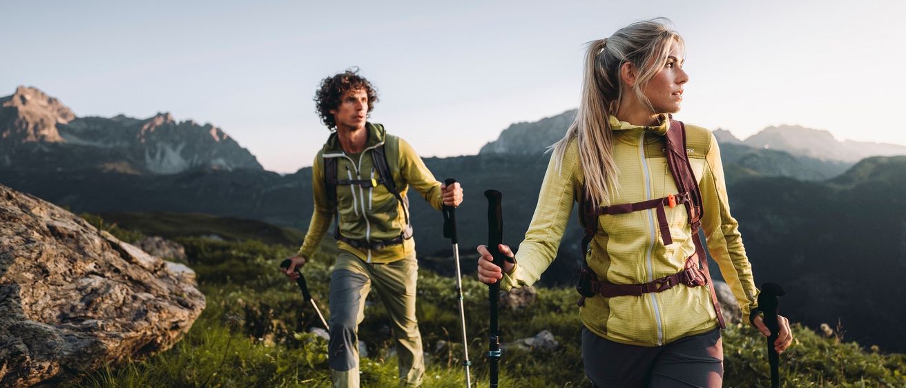 Zwei Wanderer mit Trekkingstöcken gehen auf einem grasigen Berghang. Die Frau vorne hat einen Pferdeschwanz und der Mann dahinter hat lockige Haare. Sie tragen gelbe Jacken und Hosen. Im Hintergrund sind Berge und ein klarer Himmel zu sehen.