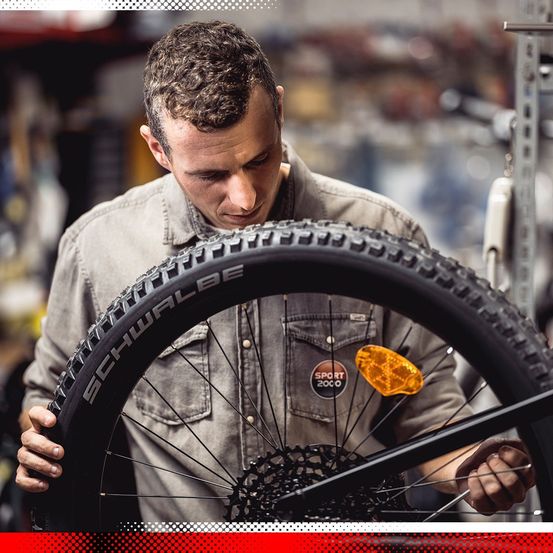 Ein Mann untersucht in einer Werkstatt ein Fahrradreifen genau, mit einem Reflektor, der am Hinterrad befestigt ist.