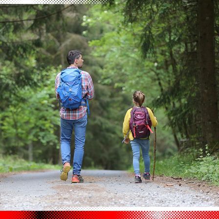 Ein Mann und ein Kind gehen auf einem Kiesweg durch einen Wald. Der Mann trägt einen Rucksack, während das Kind einen Stock und einen Rucksack hat.