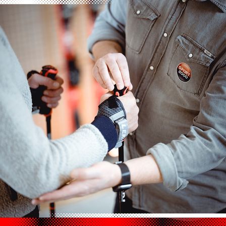 Eine Person hält einen Skistock mit einem Riemen in der Hand, während eine andere Person ihn justiert. Die Person auf der rechten Seite trägt ein graues Hemd mit einem Aufnäher.