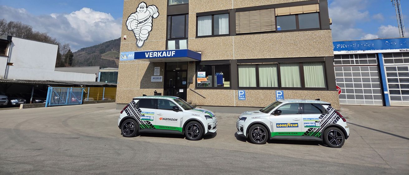 Zwei Servicewagen vor einem Edler Reifen Gebäude geparkt. Das Gebäude trägt ein großes Schild mit den Worten 'Edler Reifen' und '90 Punkte'. Ein großer Michelin-Mascot ist ebenfalls an der Wand zu sehen.