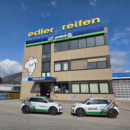 Zwei Servicefahrzeuge sind vor dem Edler Reifen-Gebäude geparkt, mit einem Schild mit Michelin-Maskottchen und der Aufschrift 'Edler Reifen 90 Points Reifen+Rader+Service'. Das Gebäude hat Glasfenster und einen blauen Vorbau.